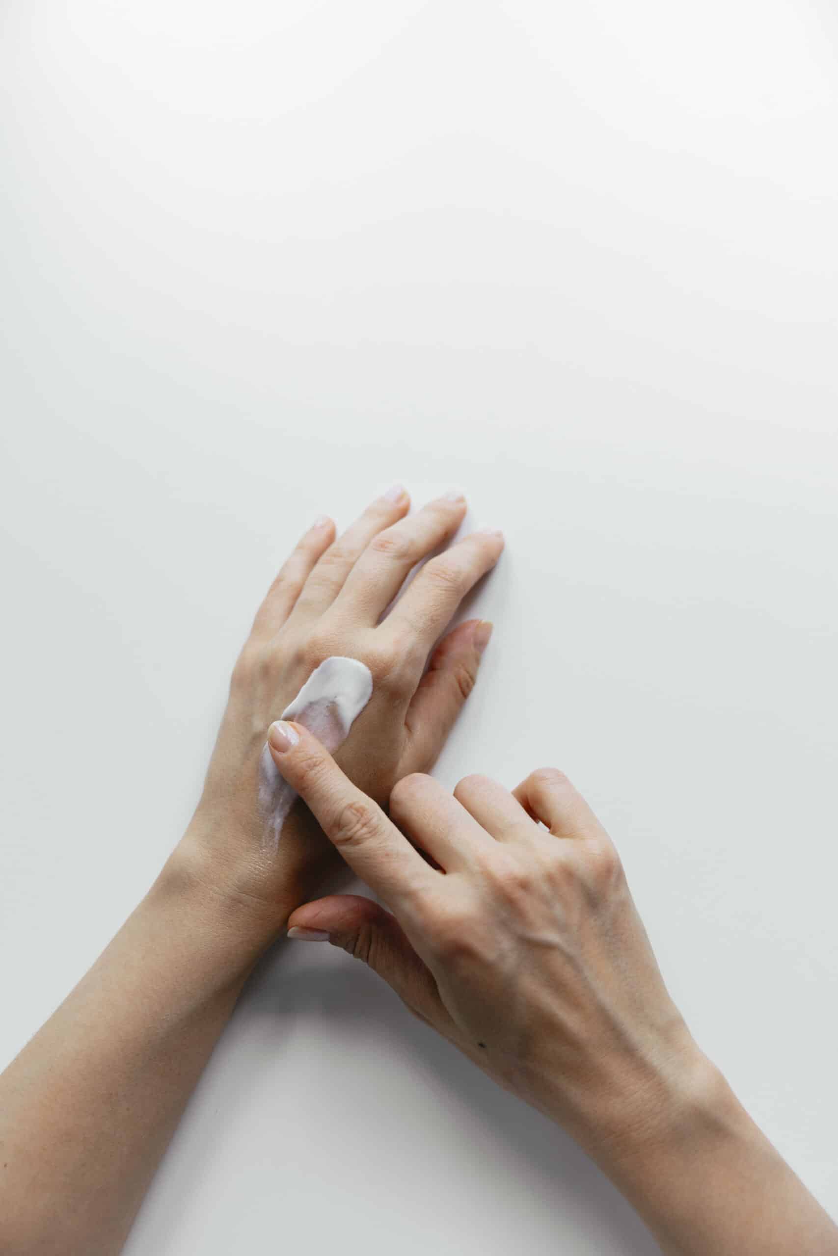 Duas mãos de mulher num fundo branco. O indicador direito espalha creme hidratatnte branco nas costas da mão esquerda